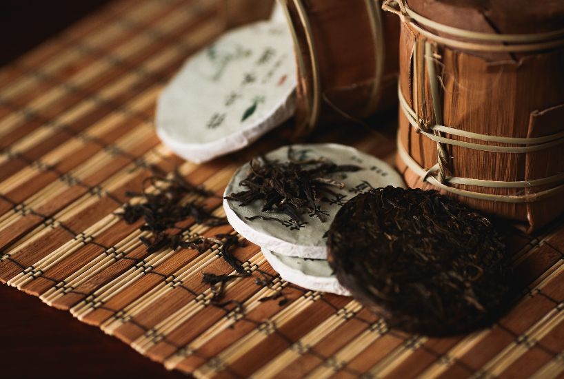 Compressed Pu-erh tea cakes with loose tea leaves on a bamboo mat, showcasing traditional Chinese tea preparation